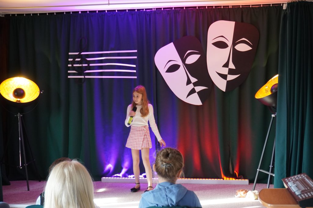 Na scenie dziewczynka w spódniczce śpiewająca. W tle dwie maski teatralne oraz pięciolinia z kluczem wiolinowym.