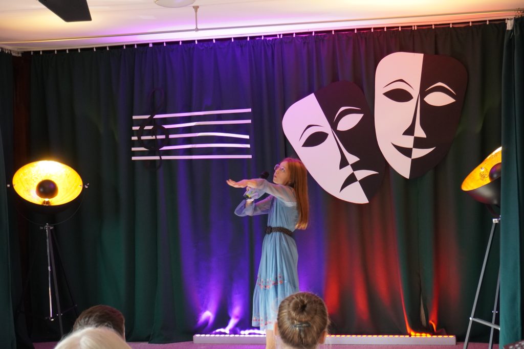 Na scenie dziewczyna śpiewająca w długiej turkusowej sukience. W tle dwie maski teatralne oraz pięciolinia z kluczem wiolinowym.